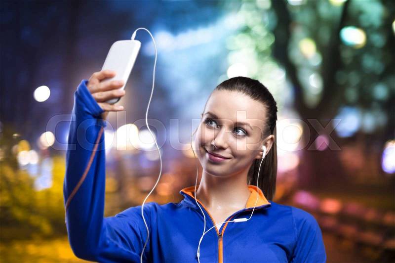 Young woman jogging at night in the city taking selfie, stock photo