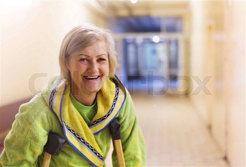 Senior woman injured sitting in the hallway of hospital holding crutches, stock photo