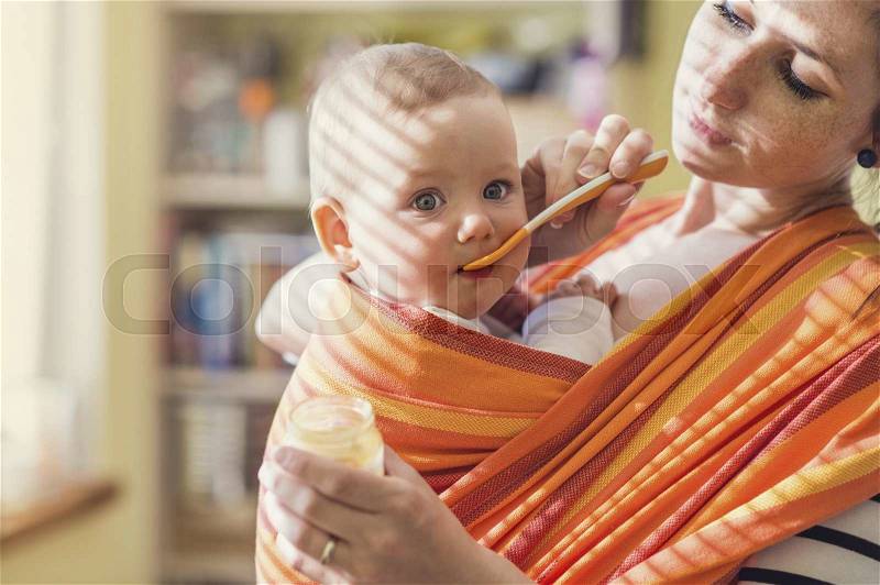 Young mother feeding her little daughter that she has in a baby carrier, stock photo
