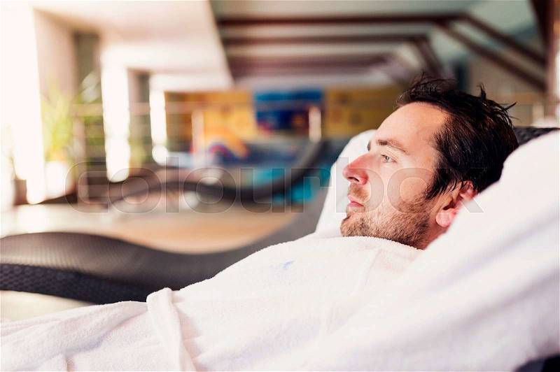 Man in white bathrobe sitting in lounge chair in spa room, stock photo
