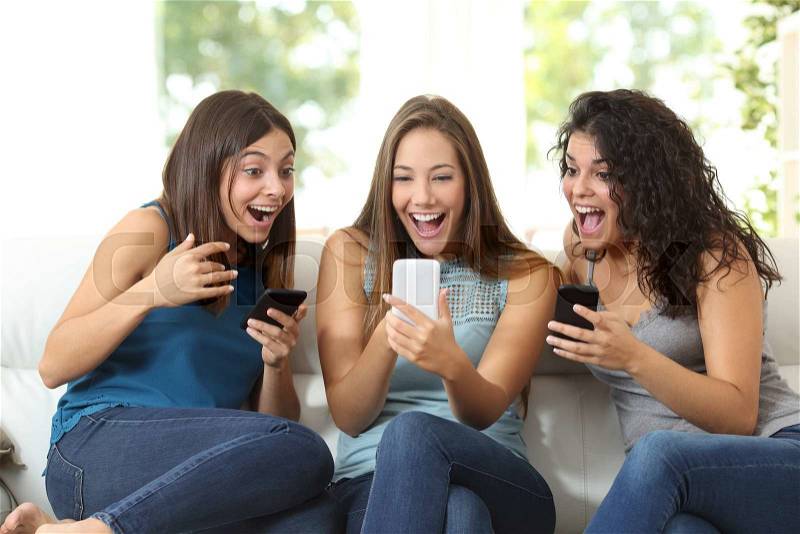 Three friends amazed watching a smart phone sitting on a couch at home, stock photo
