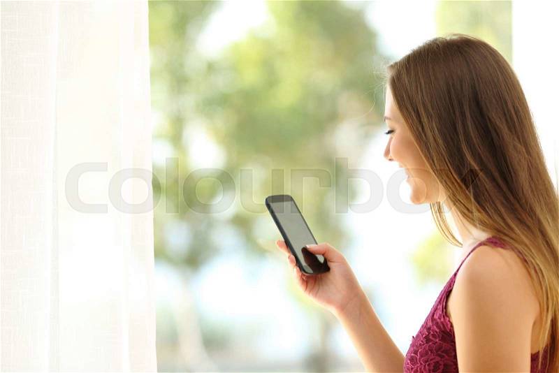 Woman waking up and using a smart phone near a bedroom window, stock photo