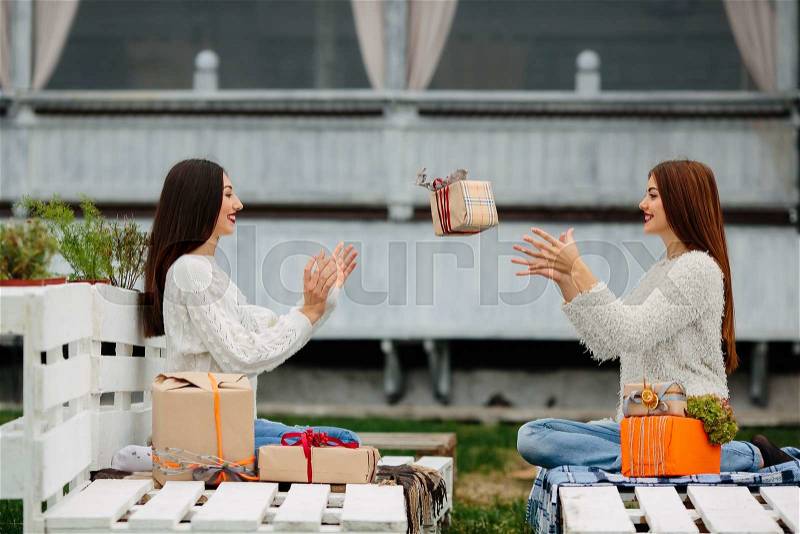 Two beautiful girls sitting on a bench and throws gifts to each other, stock photo