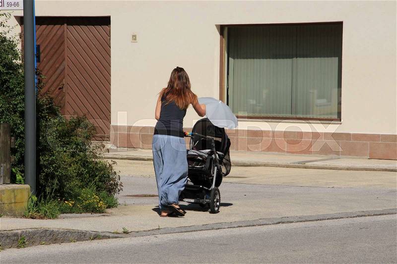 The lady walks with the pram with a blue umbrella against the sun on the pavement in the residential area in the village Rattenberg in Austria in the summer, stock photo