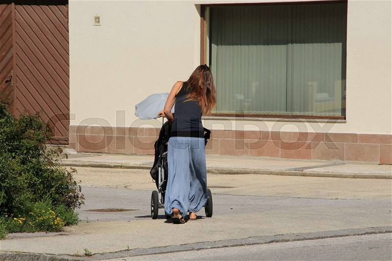 The lady walks with the pram with a blue umbrella against the sun on the pavement in the residential area in the village Rattenberg in Austria in the summer, stock photo