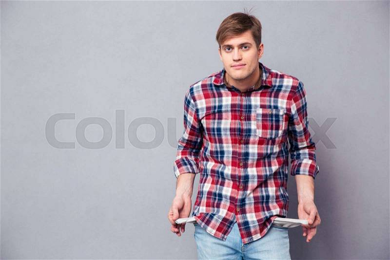 Poor handsome young man in checkered shirt and jeans showing empty pockets over grey background, stock photo