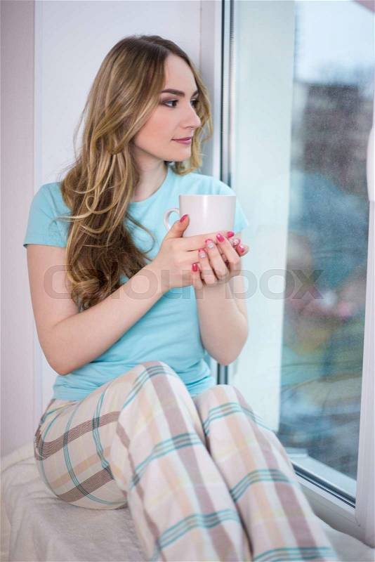 Portrait of beautiful young woman in pajamas sitting by the window with cup of tea or coffee, stock photo