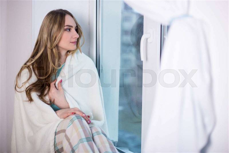 Beautiful young woman in pajamas wrapped in blanket sitting by the window, stock photo