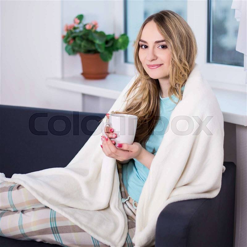 Winter concept young woman in pajamas wrapped in blanket sitting in living room with cup of tea, stock photo
