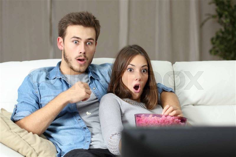 Amazed couple watching tv sitting on a couch at home, stock photo