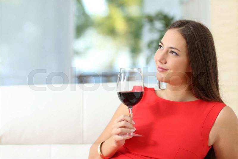 Pensive relaxed fashion woman in red with a cup of wine sitting on a couch at home, stock photo