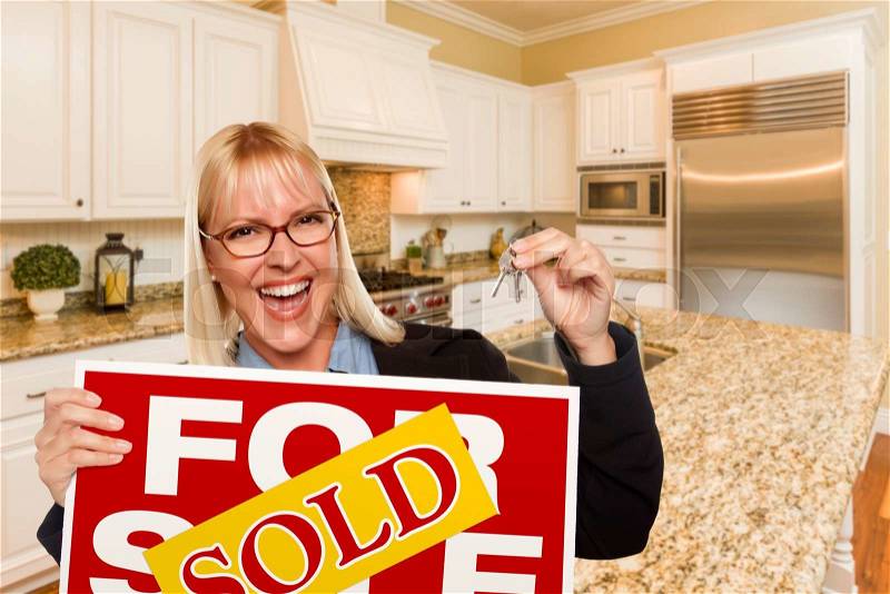 Happy Young Woman Holding Sold For Sale Real Estate Sign and Keys Inside Beautiful Custom Kitchen, stock photo