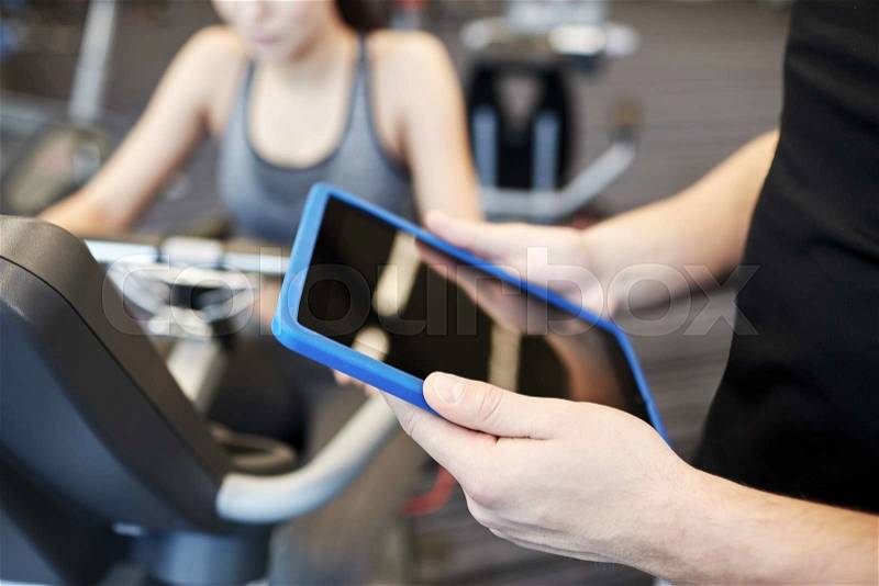 Sport, fitness, lifestyle, technology and people concept - close up of trainer hands with tablet pc computer and woman working out on exercise bike in gym, stock photo