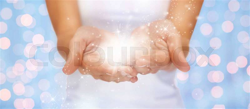 People and magic concept - close up of twinkles or fairy dust on female cupped hands over blue background with holidays lights, stock photo