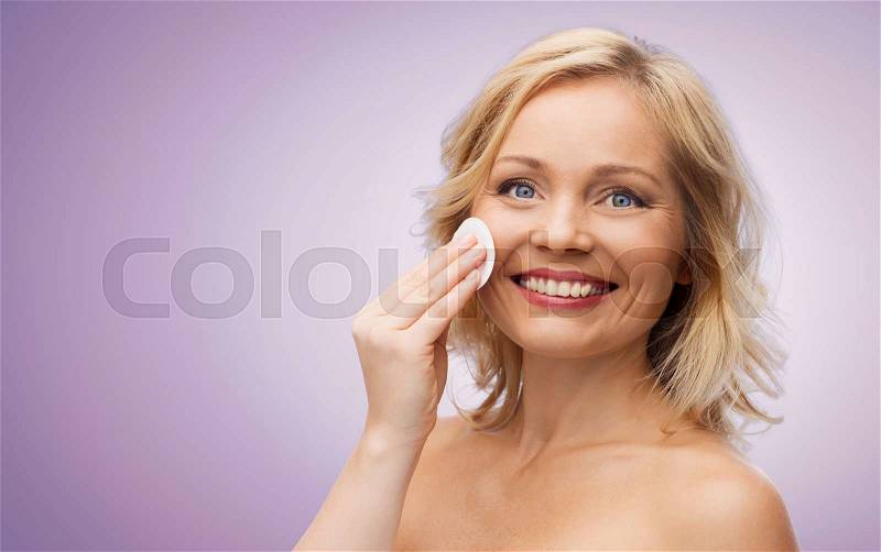 Beauty, people and skincare concept - happy middle aged woman cleaning face and removing make up with cotton pad over violet background, stock photo
