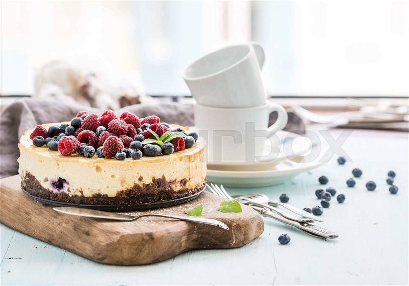 Cheesecake with fresh raspberries and blueberries on a wooden serving board, plates, cups, kitchen napkin, silverware over blue background, window at the backdrop, stock photo