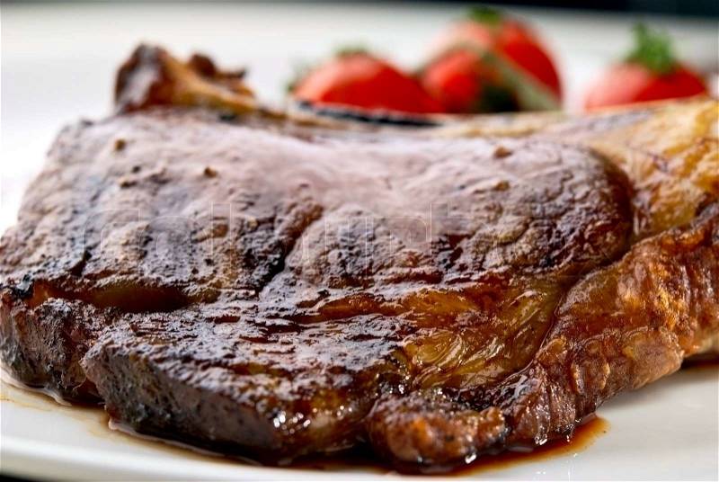 Club Steak. Veal on the bone. Macro of grilled meat ribs on white plate with cherry tomatoes and dark hot sauce, stock photo