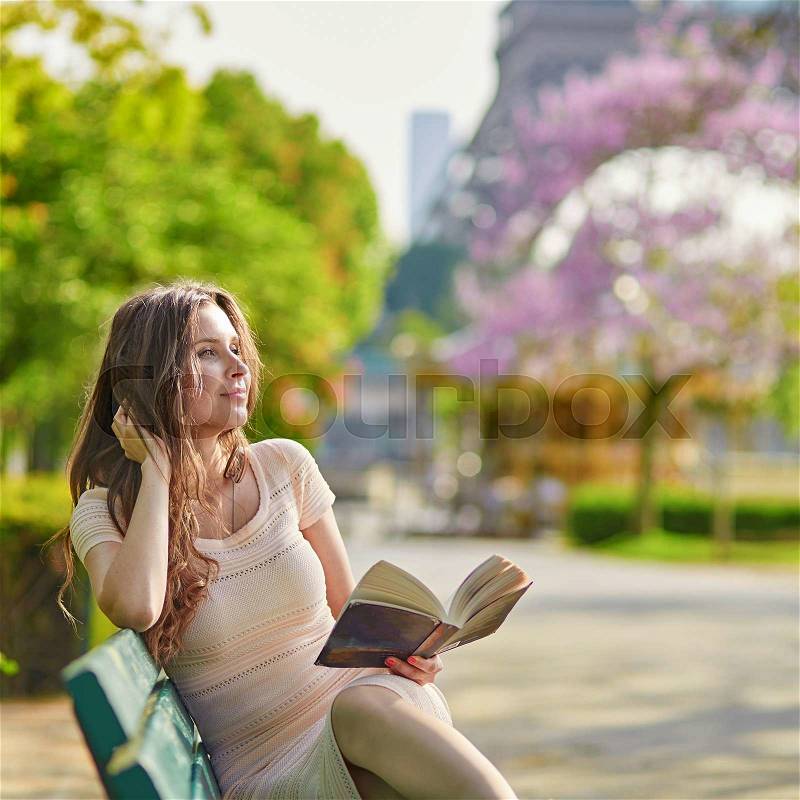 Beautiful young woman in Paris, near the Eiffel tower on a nice and sunny spring day, reading on the bench outdoors, stock photo