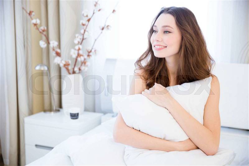 Happy charming young woman waking up in bed and hugging white pillow, stock photo