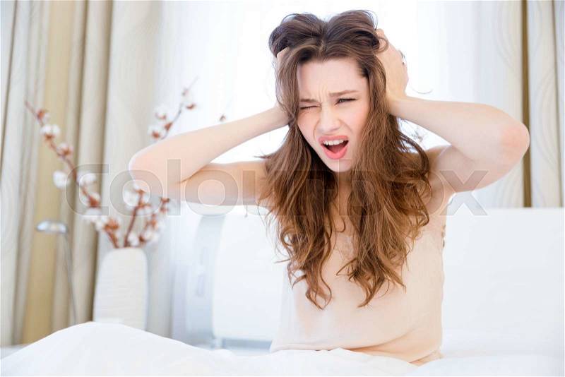 Irritated fatigued young woman sitting on bed at home with eard closed by hands , stock photo