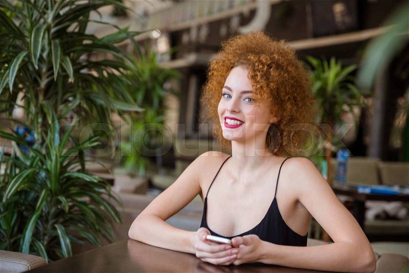 Portrait of a smiling redhead woman with curly hair sitting at the table in restaurant and looking at camera, stock photo
