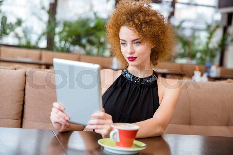 Happy redhead attractive woman with curly hair using tablet computer in restaurant, stock photo