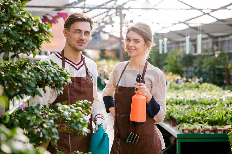 Happy man and woman gardeners holding watering can and pulveriser for spraying flowers and plants in greenhouse , stock photo