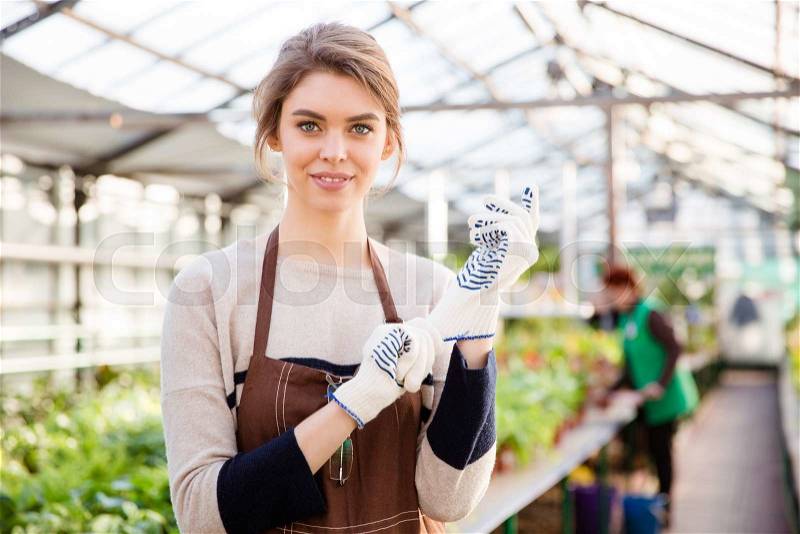 Happy young woman gardener in gerden gloves and brown apron standing in greenhouse, stock photo