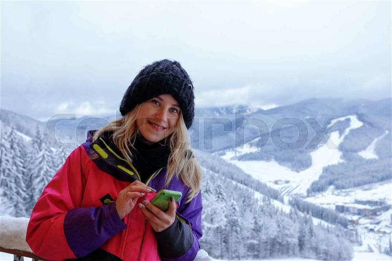 Portrait of smiling woman writes a message on the phone in the mountains. Girl in pink ski suit and blach wool hat. On the background snow-capped mountains and the forests, stock photo