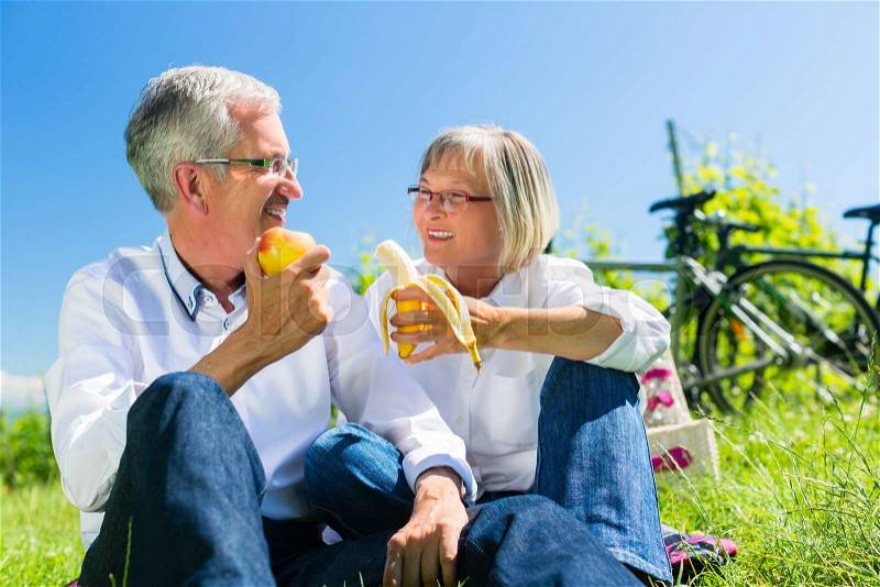 Senior couple eating fruit and drinking at picnic in summer, beautiful landscape in the background, stock photo