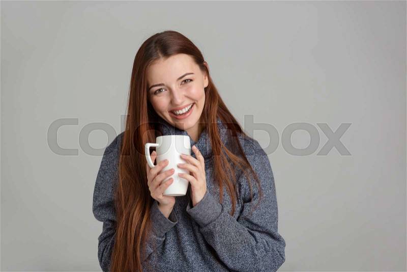 Happy attractive young woman with long hair in grey sweatshirt holding white mug and laughing over grey background, stock photo