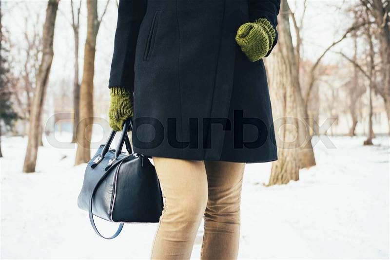Woman in a black coat, green gloves and a bag walks in the park in winter, stock photo