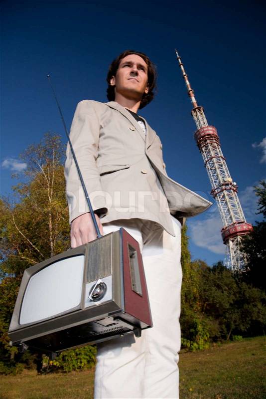 Young man holding portable retro tv set in front of high television tower, stock photo