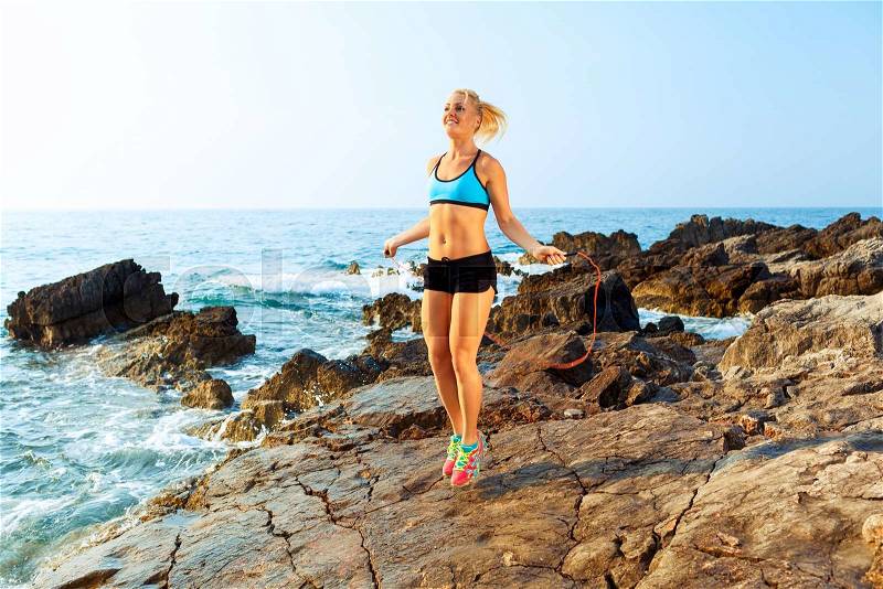 Beautiful athlete woman doing sport exercising with the rope on the rocks by the sea, stock photo
