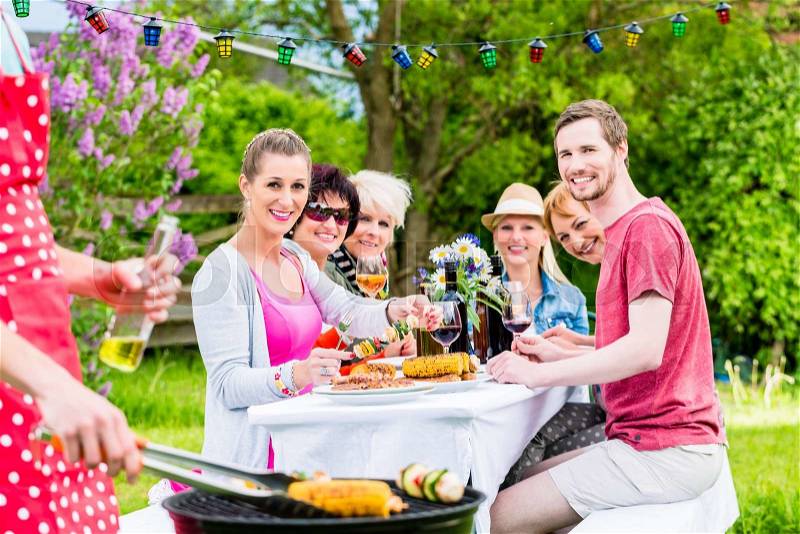 Man grilling meat and vegetables on garden party, his friends eating the bbq meat, stock photo