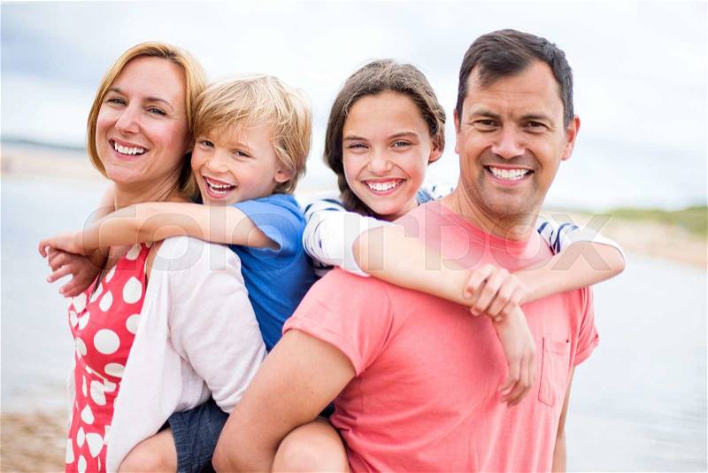 A family of four are at the beach together, the children are on their parents backs. They are all smiling looking at the camera, stock photo