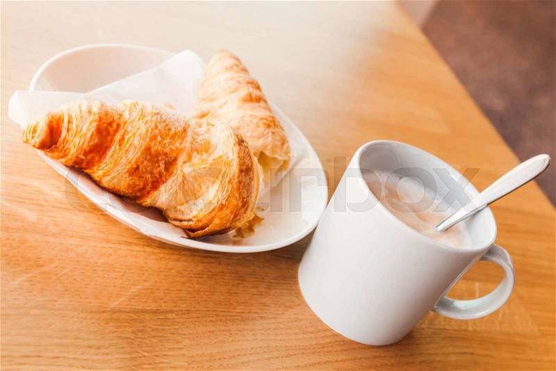 Cappuccino and croissant. Cup of coffee with milk foam stands on wooden table in cafeteria, closeup photo with selective focus, stock photo