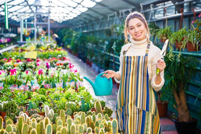 Happy harming young woman gardenet in striped apron standing and holding garden shovel and blue watering can in greenhouse, stock photo