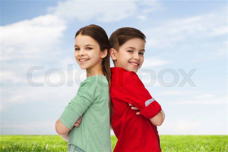 Childhood, summer and people concept - happy smiling boy and girl standing back to back over blue sky and grass background, stock photo