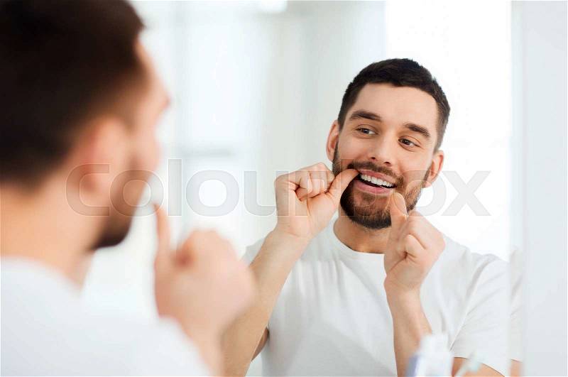 Health care, dental hygiene, people and beauty concept - smiling young man with floss cleaning teeth and looking to mirror at home bathroom, stock photo
