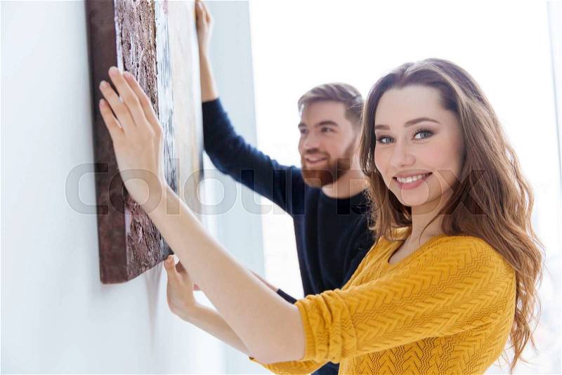 Cheerful couple hanging picture on the wall at home, stock photo