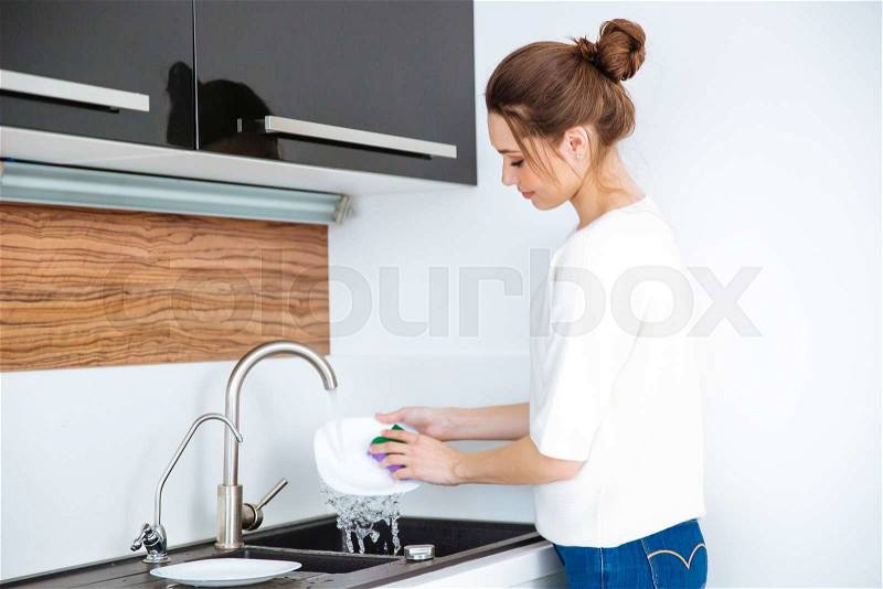 Cute lovely young woman standing and washing dishes at home, stock photo