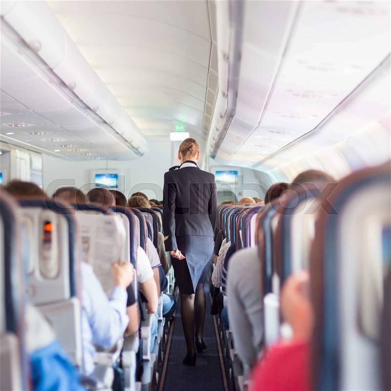 Interior of commercial airplane with passengers on seats during flight. Stewardess in dark blue uniform walking the aisle. Square composition, stock photo