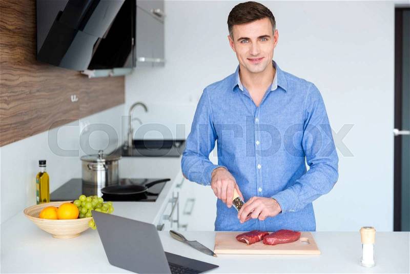 Attractive smiling young man in blue shit cooking meat on the kitchen at home, stock photo
