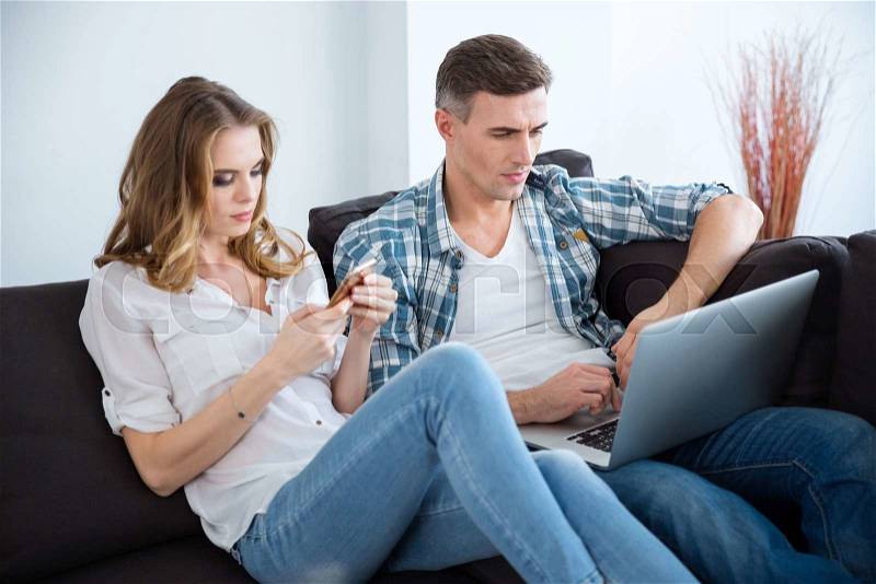 Beautiful couple sitting on sofa and using laptop and smartphone separately , stock photo