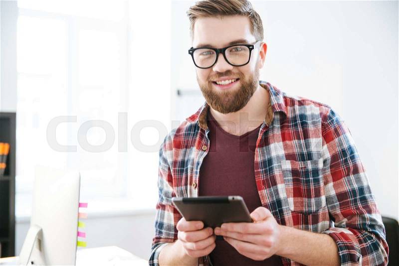 Happy handsome bearded male in glasses using tablet in the office, stock photo
