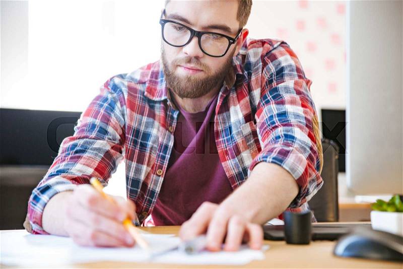 Handsome serious man in checkered shirt and glasses sitting and writing in the office, stock photo