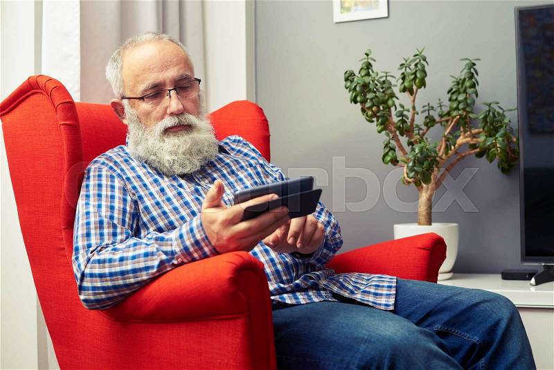 Senior man sitting on red chair and using his smartphone at home, stock photo