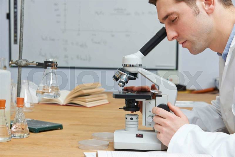 Male Biology Teacher Looking Through Microscope In Classroom, stock photo