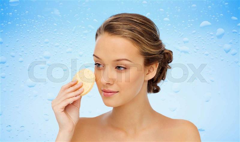 Beauty, people, moisturizing, skin care and skincare concept - young woman cleaning face with exfoliating sponge over water drops on blue background, stock photo